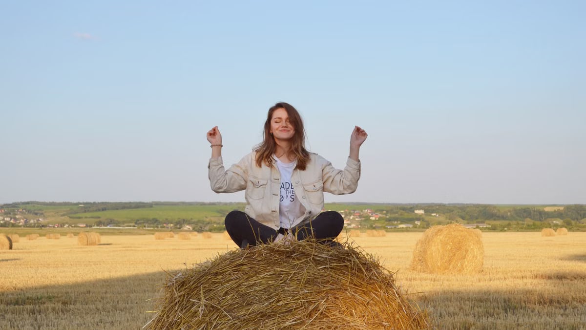 A young woman sits peacefully on a hay bale in a serene summer field.